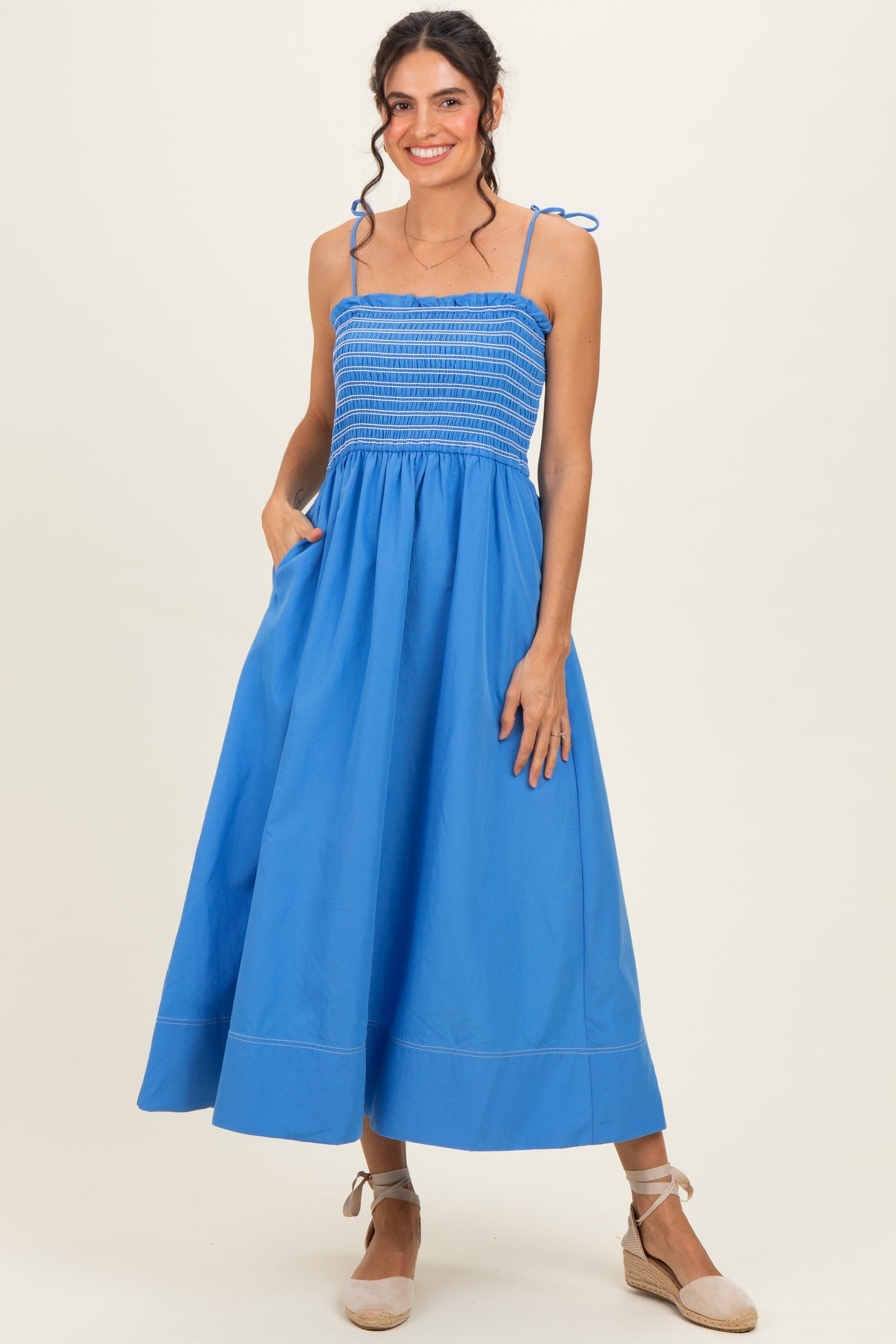 woman wearing a blue smocked maxi dress with thin straps and pockets, standing gracefully with a slight smile