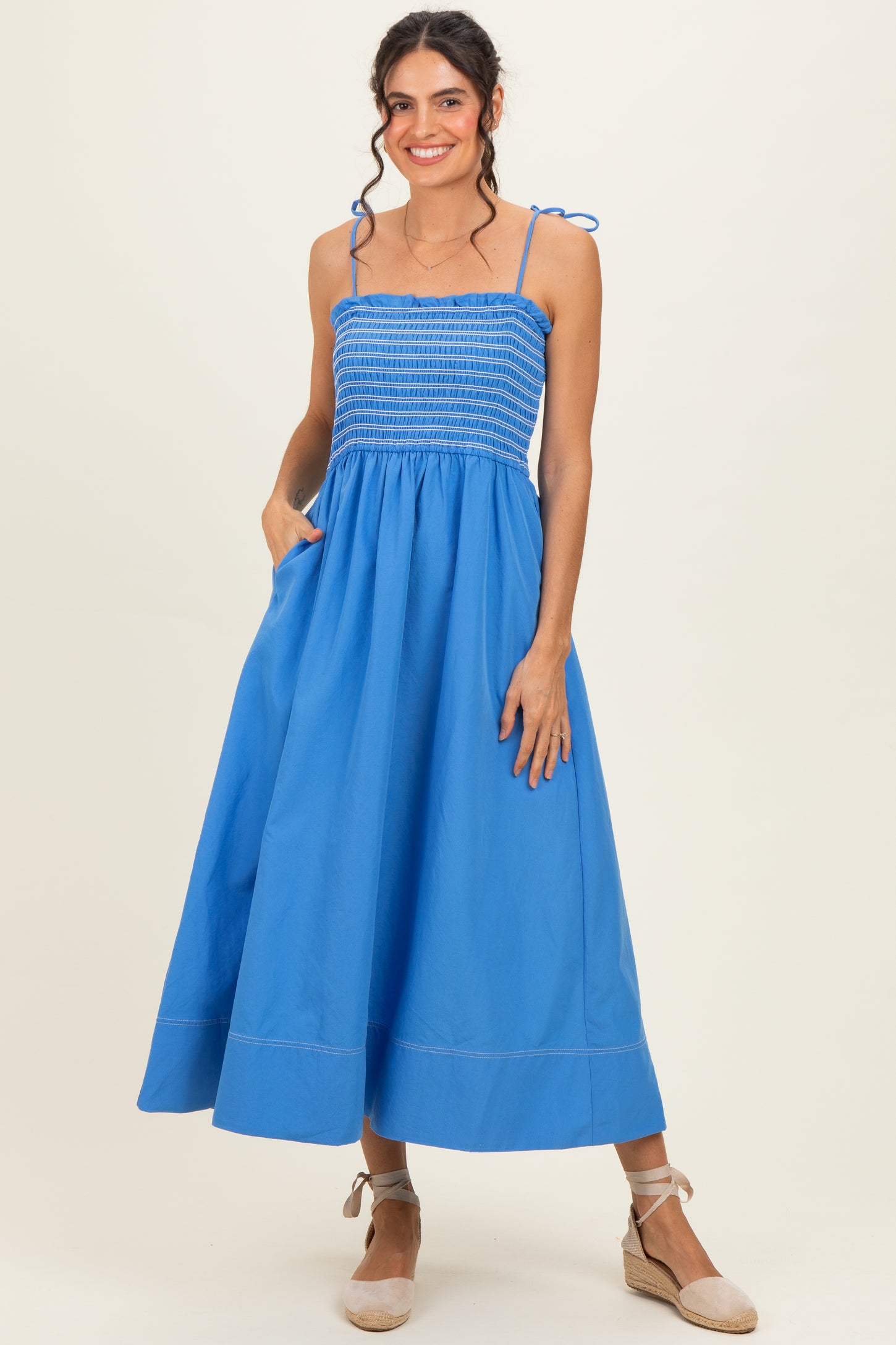 woman wearing a blue smocked maxi dress with thin straps and pockets, standing gracefully with a slight smile