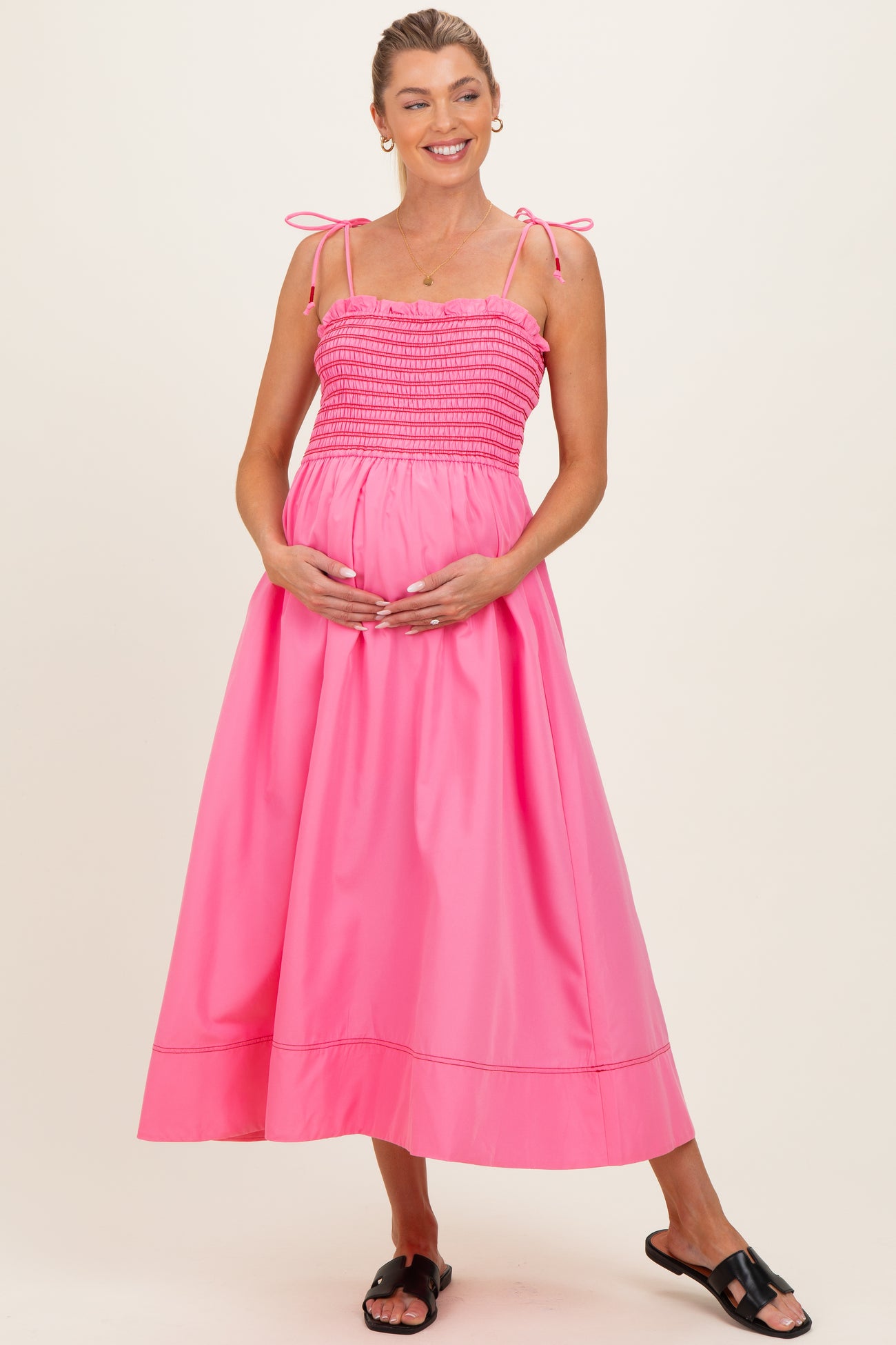 woman wearing a bright pink maternity maxi dress with spaghetti straps and a gathered bodice, holding her baby bump