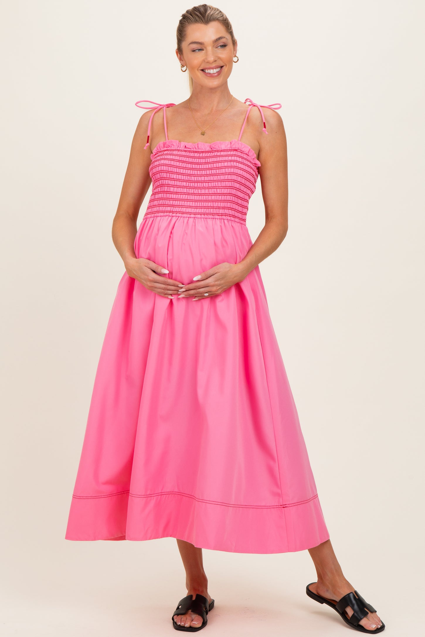 woman wearing a bright pink maternity maxi dress with spaghetti straps and a gathered bodice, holding her baby bump
