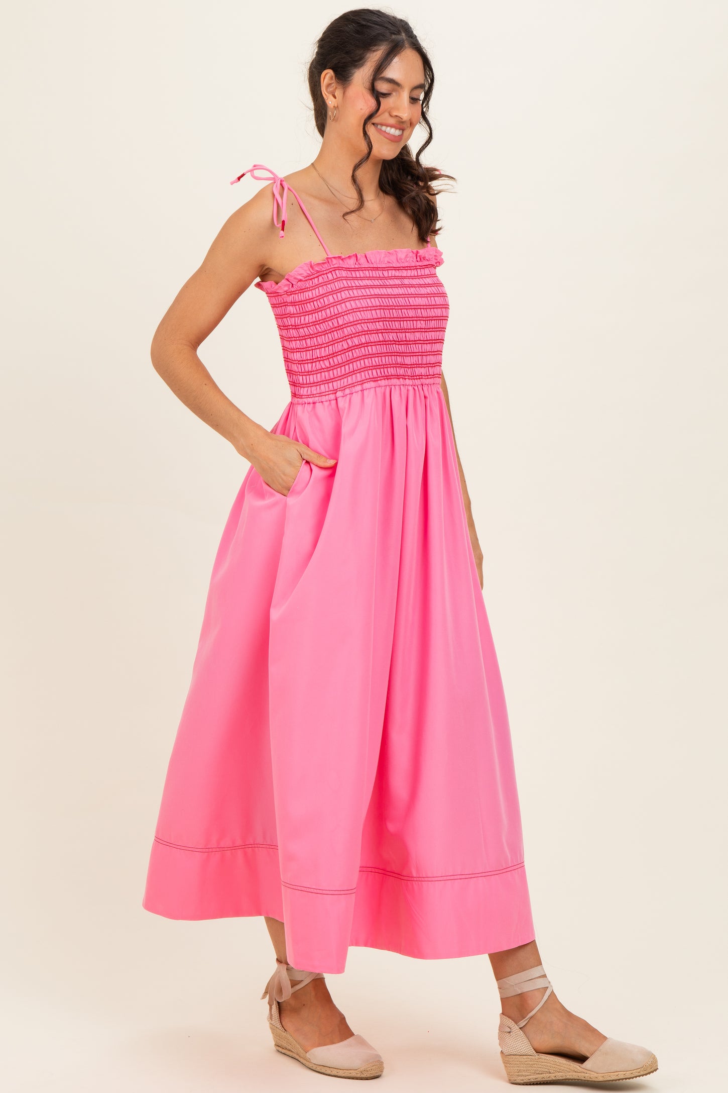 woman wearing a bright pink maxi dress with smocked bodice and tie straps, featuring side pockets and a flowy skirt