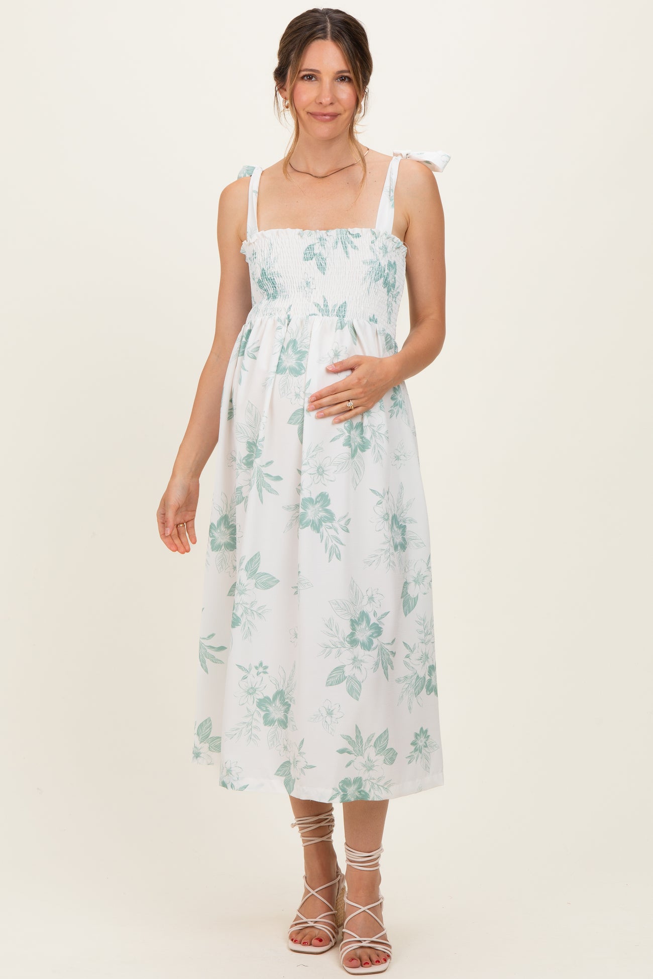 woman wearing a white floral maternity dress with smocked bodice and tie straps, gently holding her baby bump