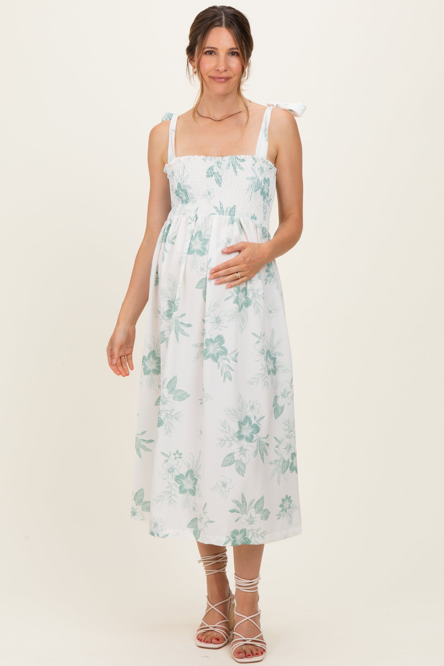 woman wearing a white floral maternity dress with smocked bodice and tie straps, gently holding her baby bump