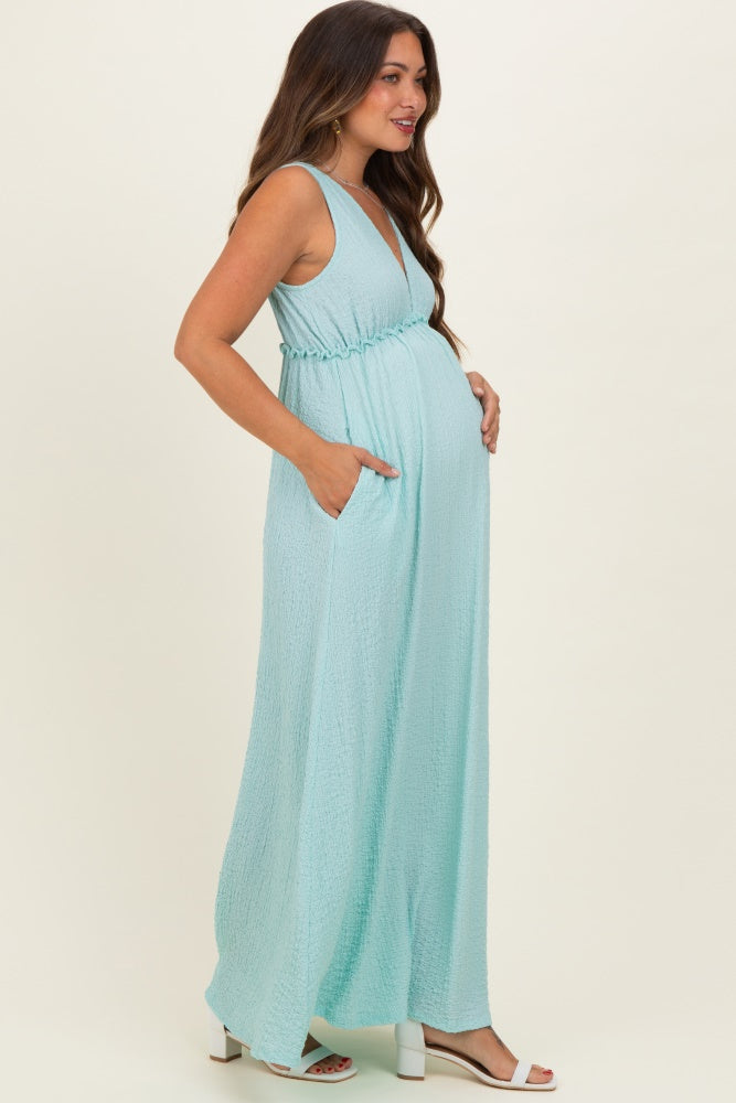 woman wearing a light blue sleeveless maternity maxi dress with a tie waist and pockets, showcasing her baby bump