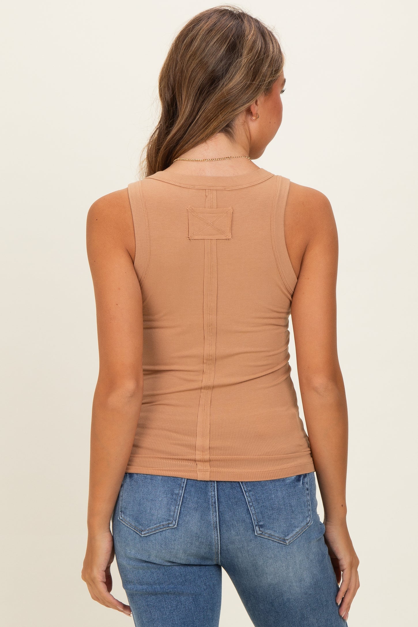 woman wearing a beige sleeveless fitted top with a low back detail, paired with blue jeans, viewed from the back