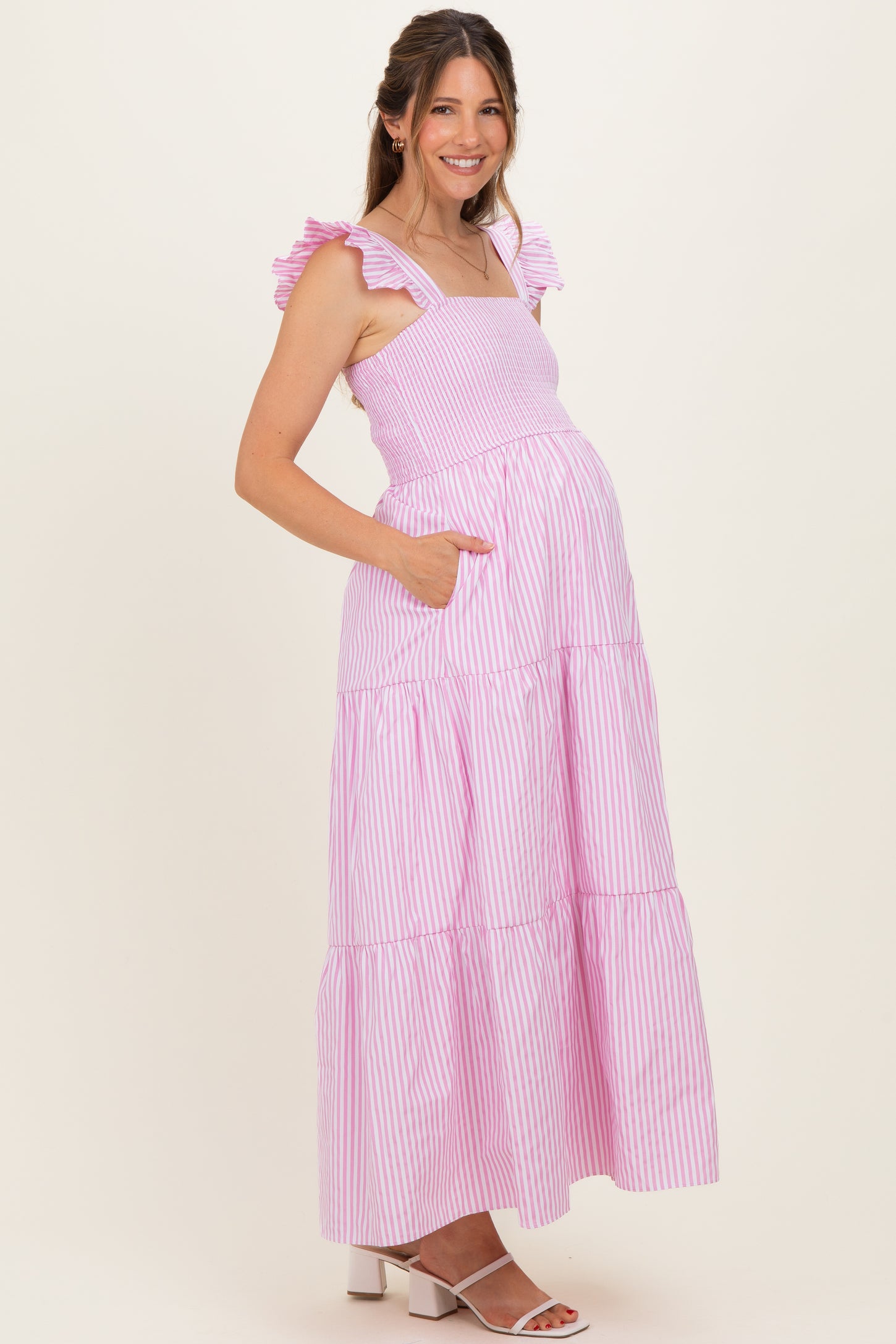 woman wearing a pink and white striped maternity maxi dress with ruffle straps and pockets, showcasing her baby bump