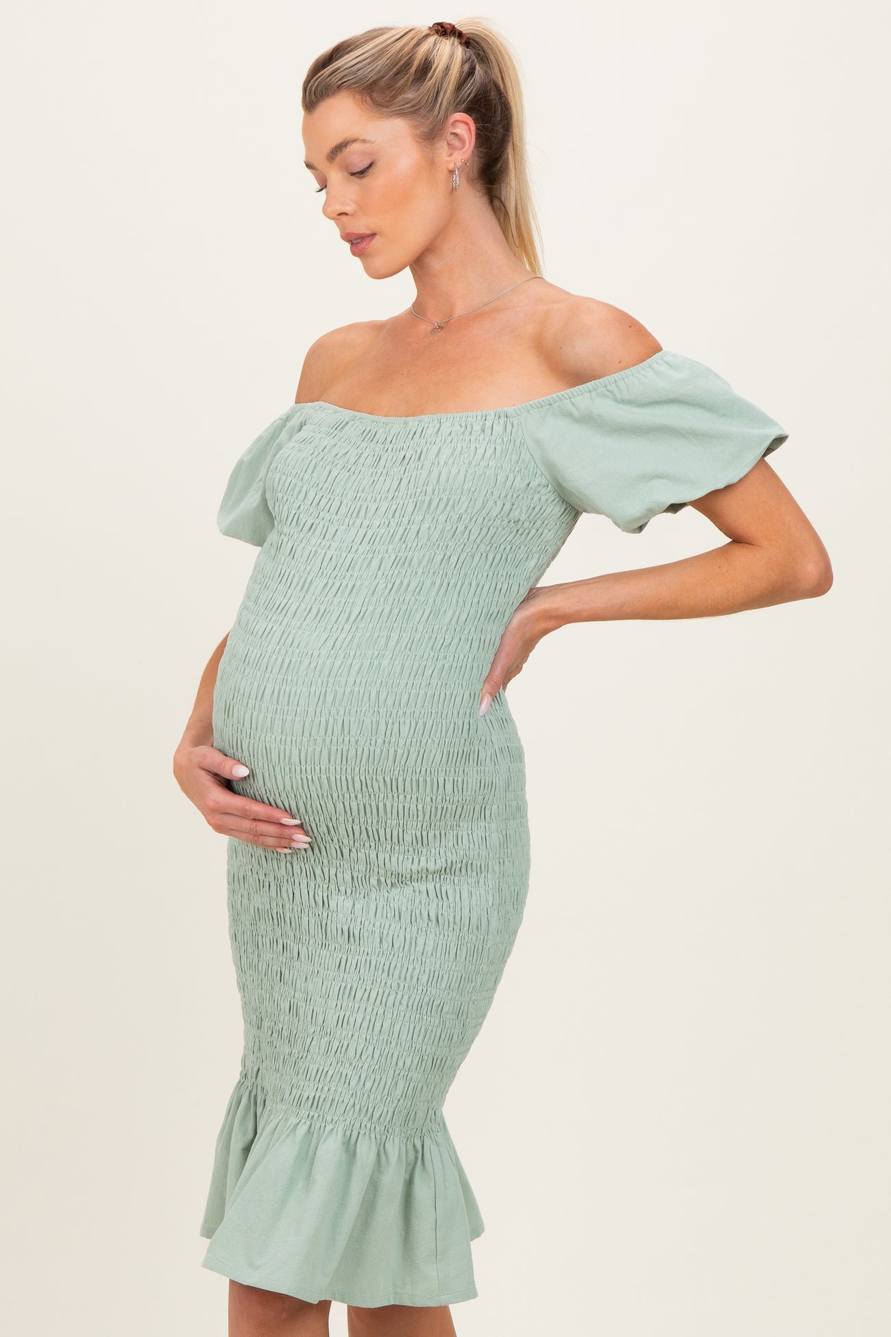 woman wearing a light green off-the-shoulder maternity dress with a fitted silhouette and ruffled hem, cradling her baby bump