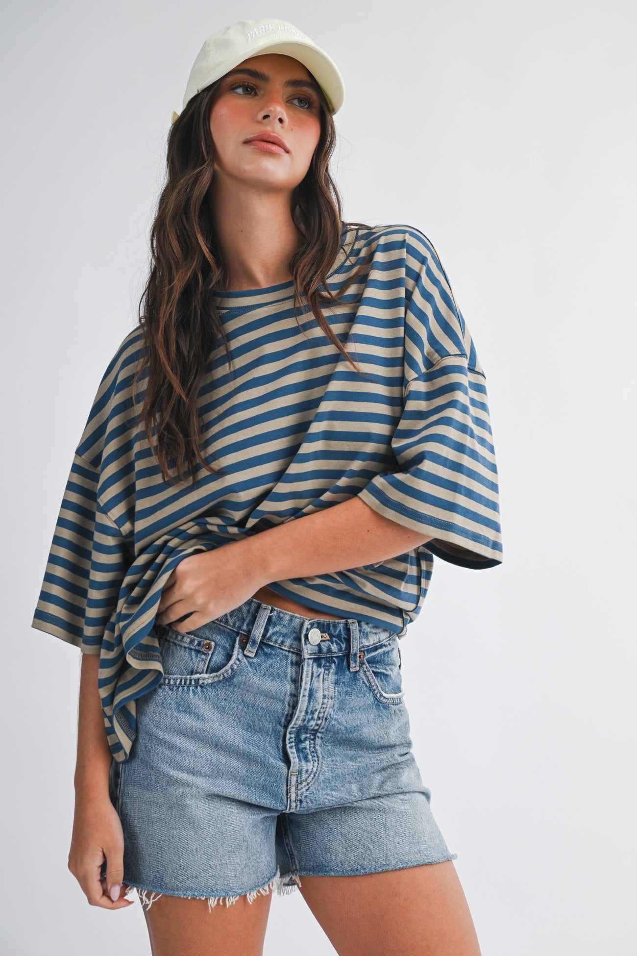 woman wearing a striped oversized t-shirt with rolled sleeves and denim shorts, accessorized with a light green cap