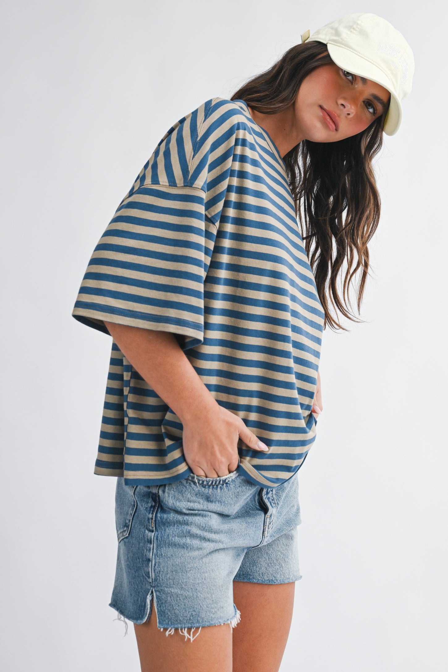 woman wearing a blue and beige striped oversized t-shirt with rolled sleeves and denim shorts, accessorized with a white cap