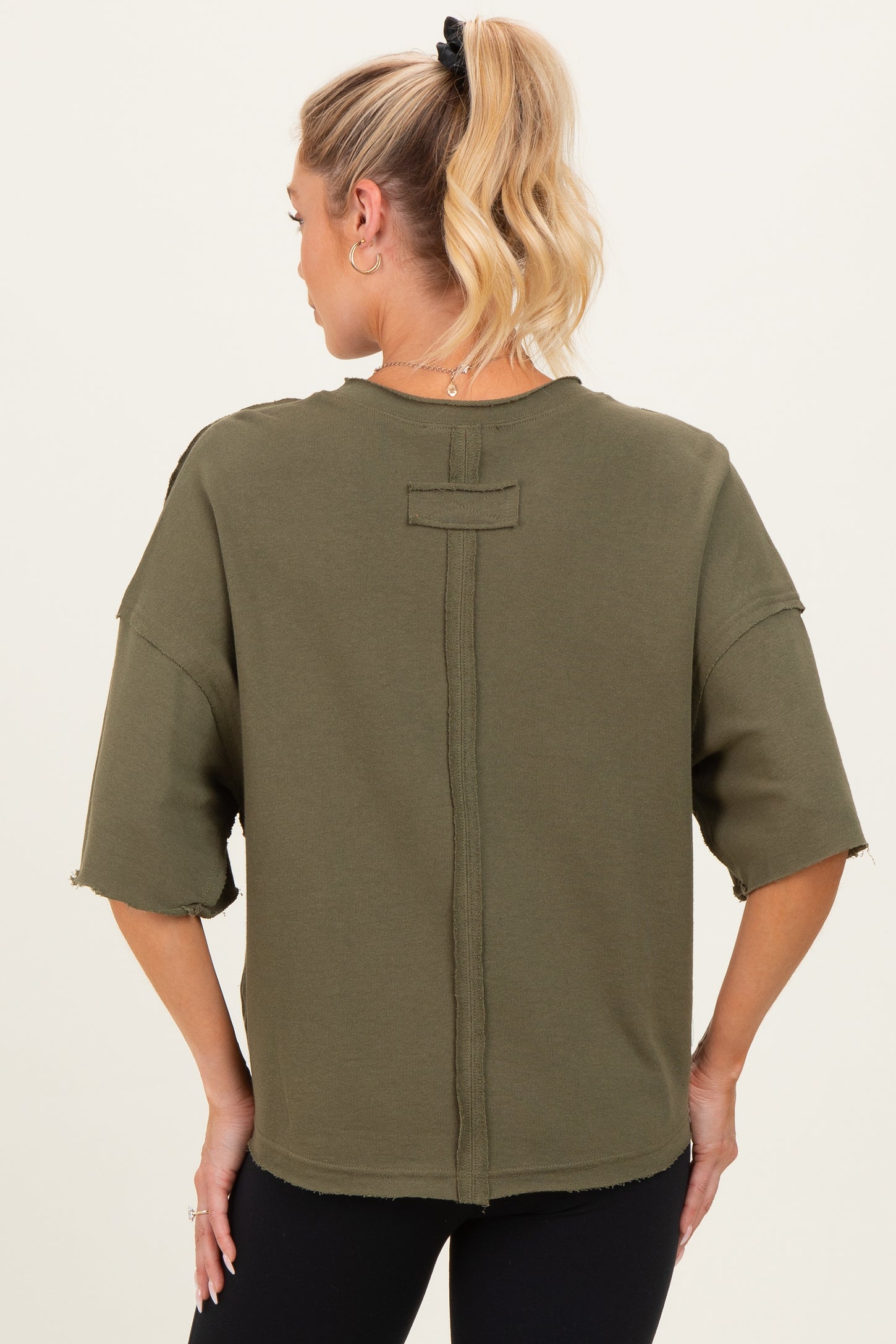 woman wearing an olive green maternity top with a relaxed fit and raw hem details, showcasing the back style
