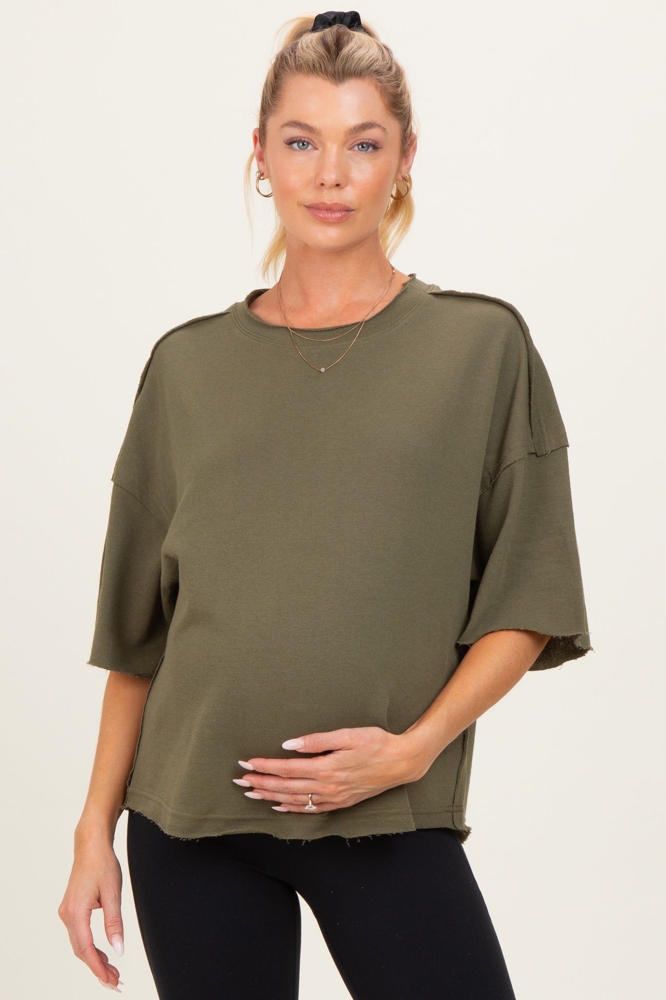 woman wearing an olive green oversized maternity top with a textured hem and holding her baby bump
