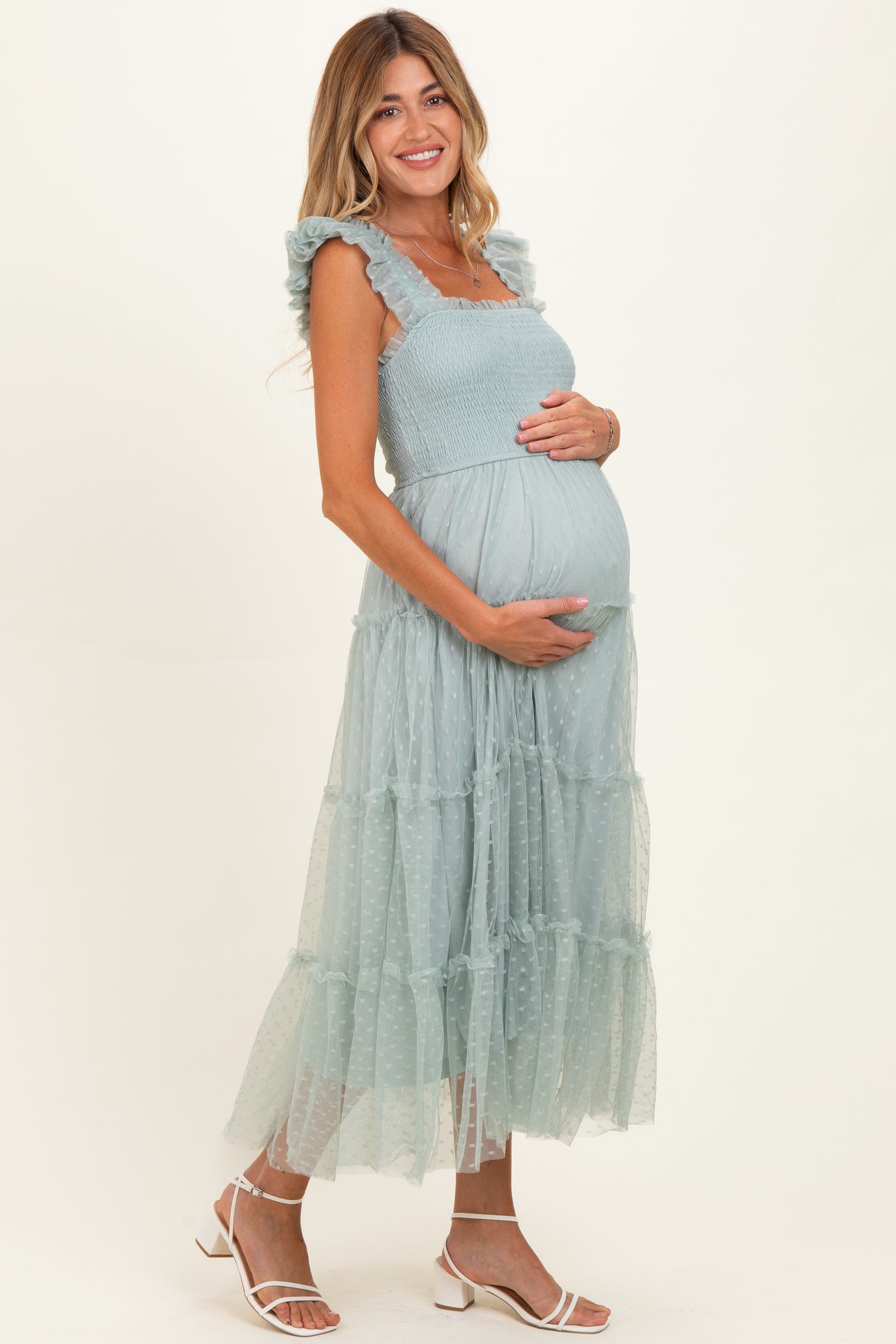 woman wearing a light blue maternity dress with ruffled straps and tiered tulle skirt, cradling her baby bump