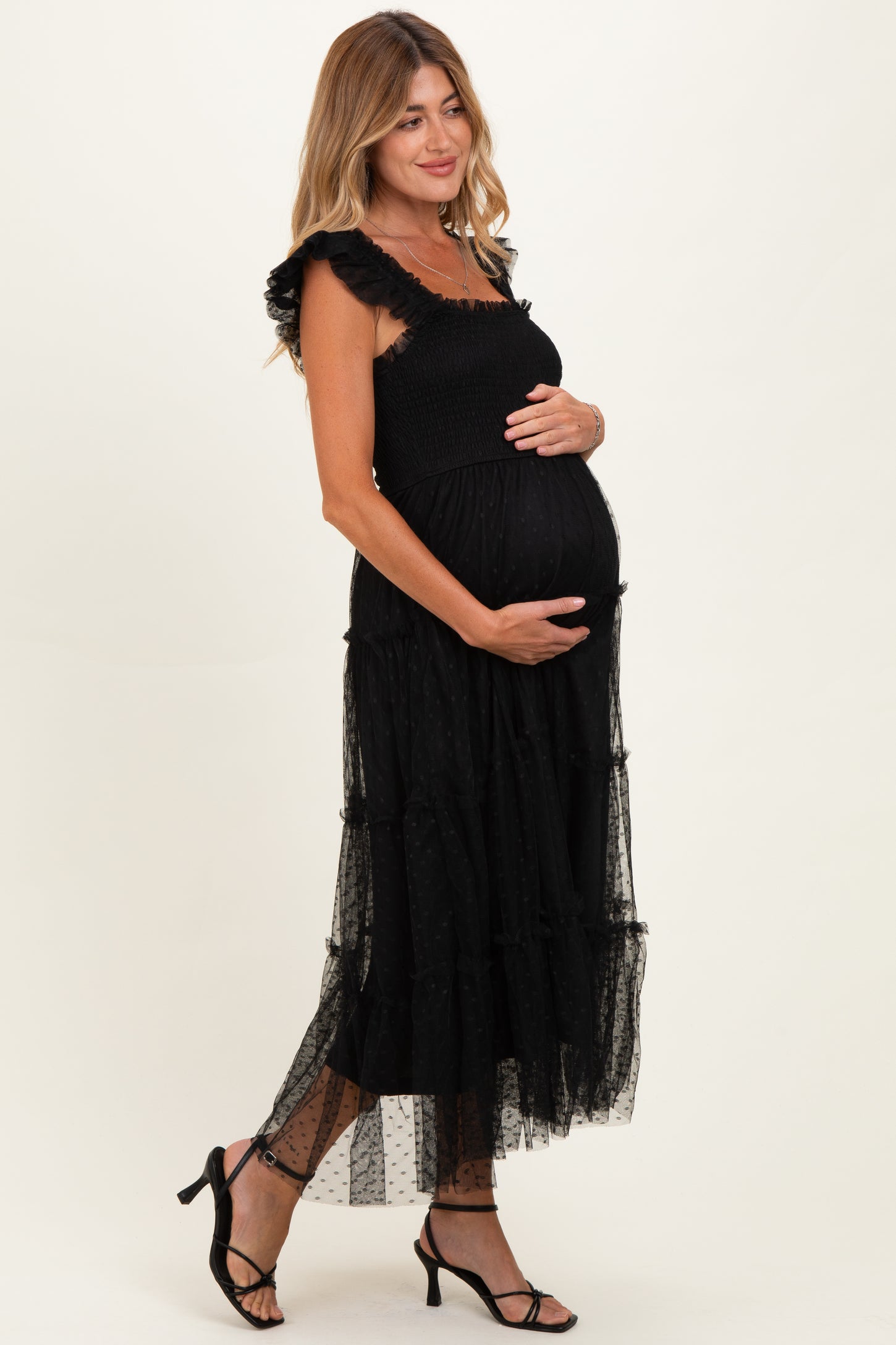woman wearing a black polka dot maternity maxi dress with ruffled straps and crinkled tiers, holding her baby bump