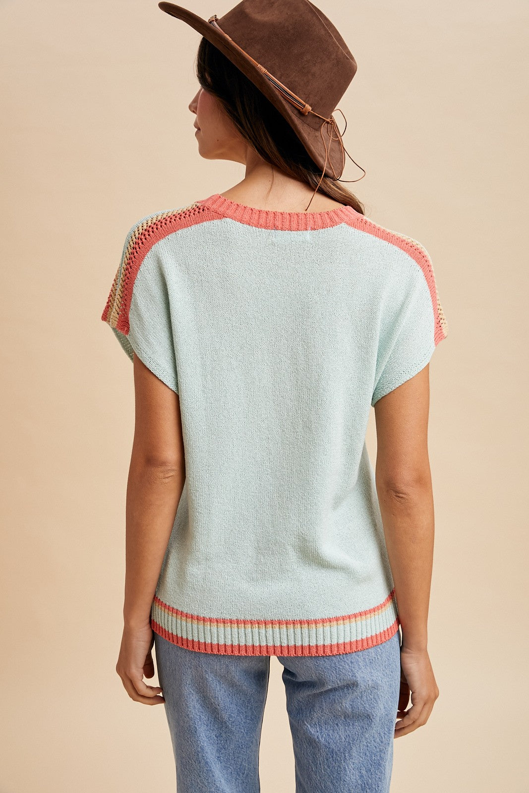 woman wearing a mint green short sleeve sweater with coral accents and back detail, paired with light blue jeans and a brown hat