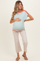 woman wearing a light blue maternity top with coral accents and cream cropped pants, holding her baby bump