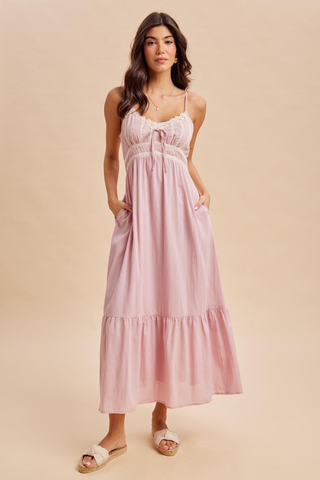 woman wearing a light pink maxi dress with adjustable straps, lace detailing, and pockets