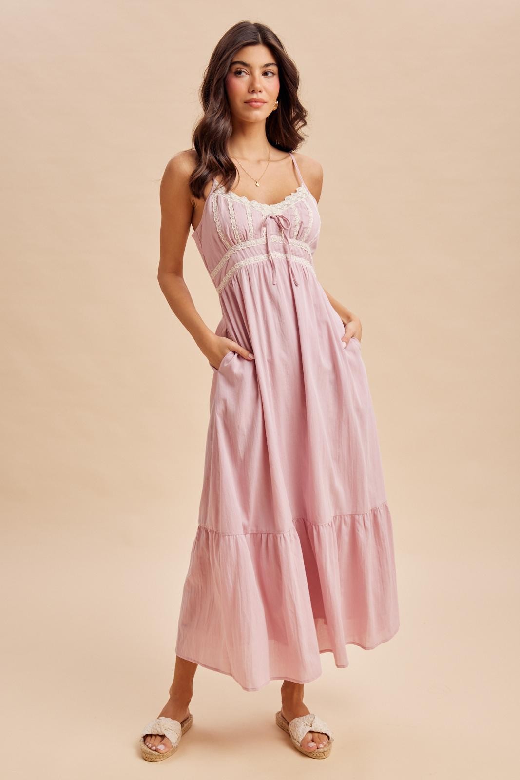 woman wearing a light pink maxi dress with lace detail and pockets, standing against a neutral background