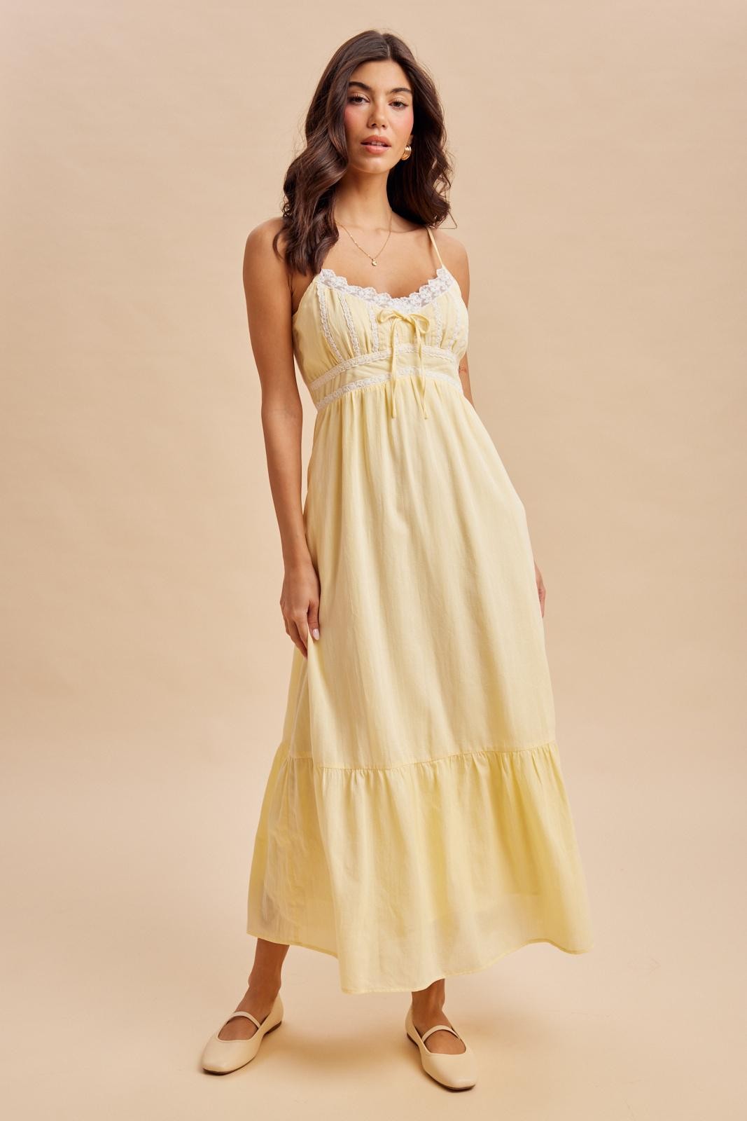 woman wearing a light yellow maxi dress with lace details and a tiered skirt, standing on a neutral background