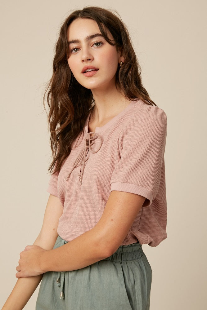 woman wearing a pink short sleeve top with a lace-up front detail and styled with light green pants