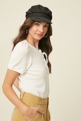 woman wearing a white ribbed short sleeve blouse with a lace-up neckline and a gray newsboy cap