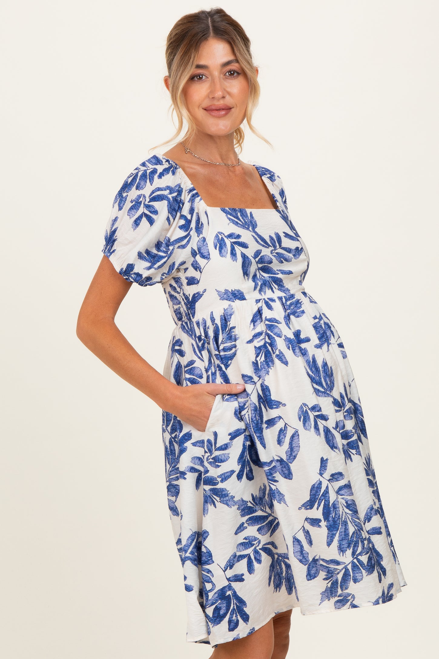 woman wearing a white short sleeve maternity dress with blue floral print and pockets, showcasing her baby bump
