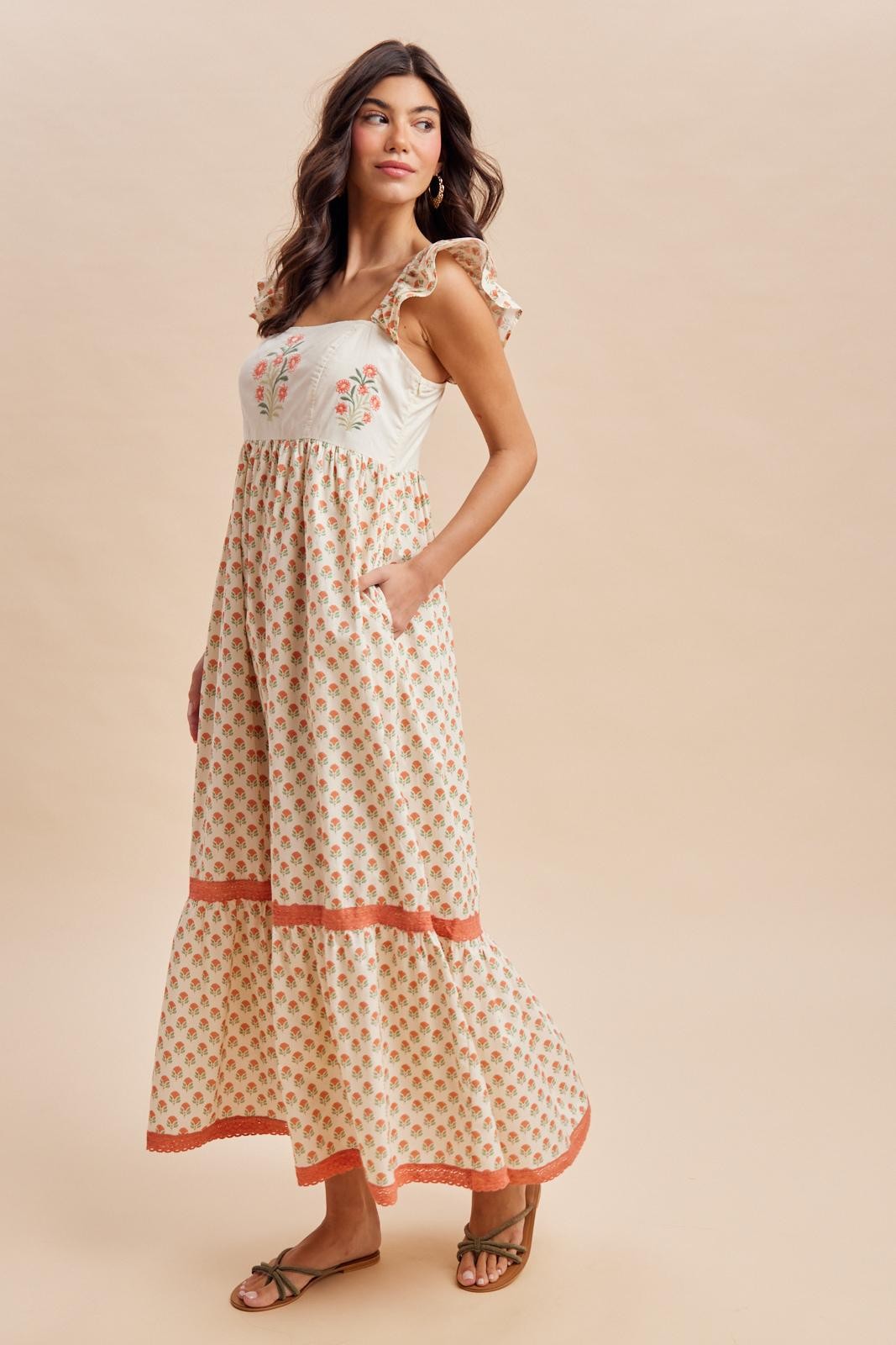 woman wearing a cream maxi dress with floral embroidery, ruffle straps, and pockets, styled with flat sandals
