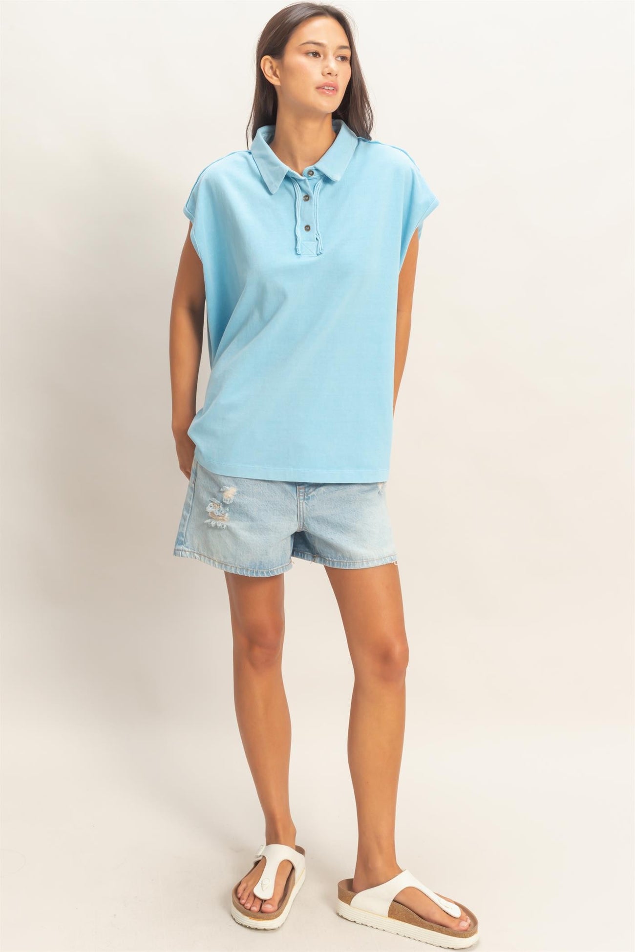 woman wearing a light blue short sleeve polo shirt with buttons and distressed denim shorts, paired with white slide sandals