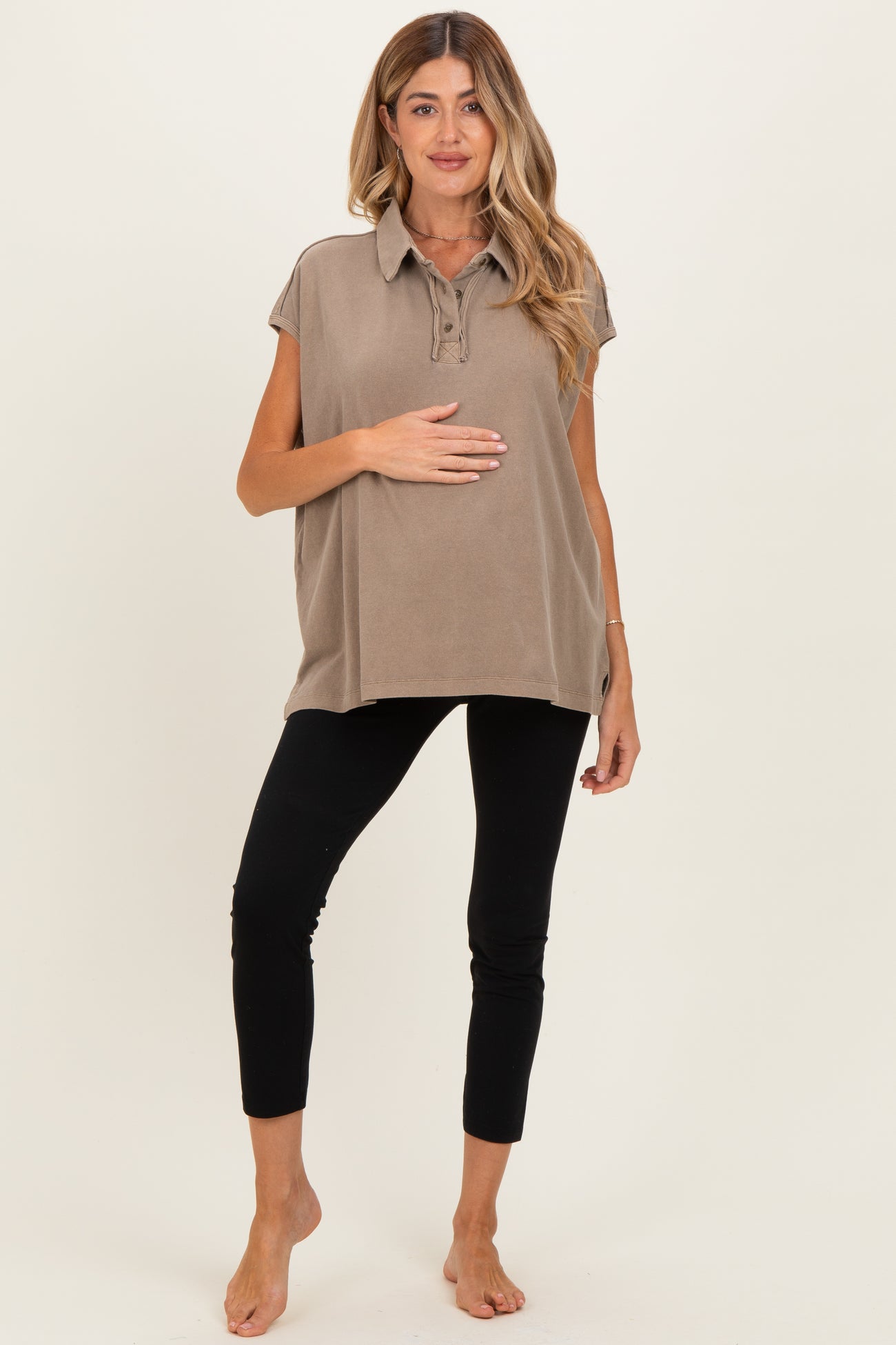 woman wearing a taupe maternity polo shirt and black leggings, holding her baby bump