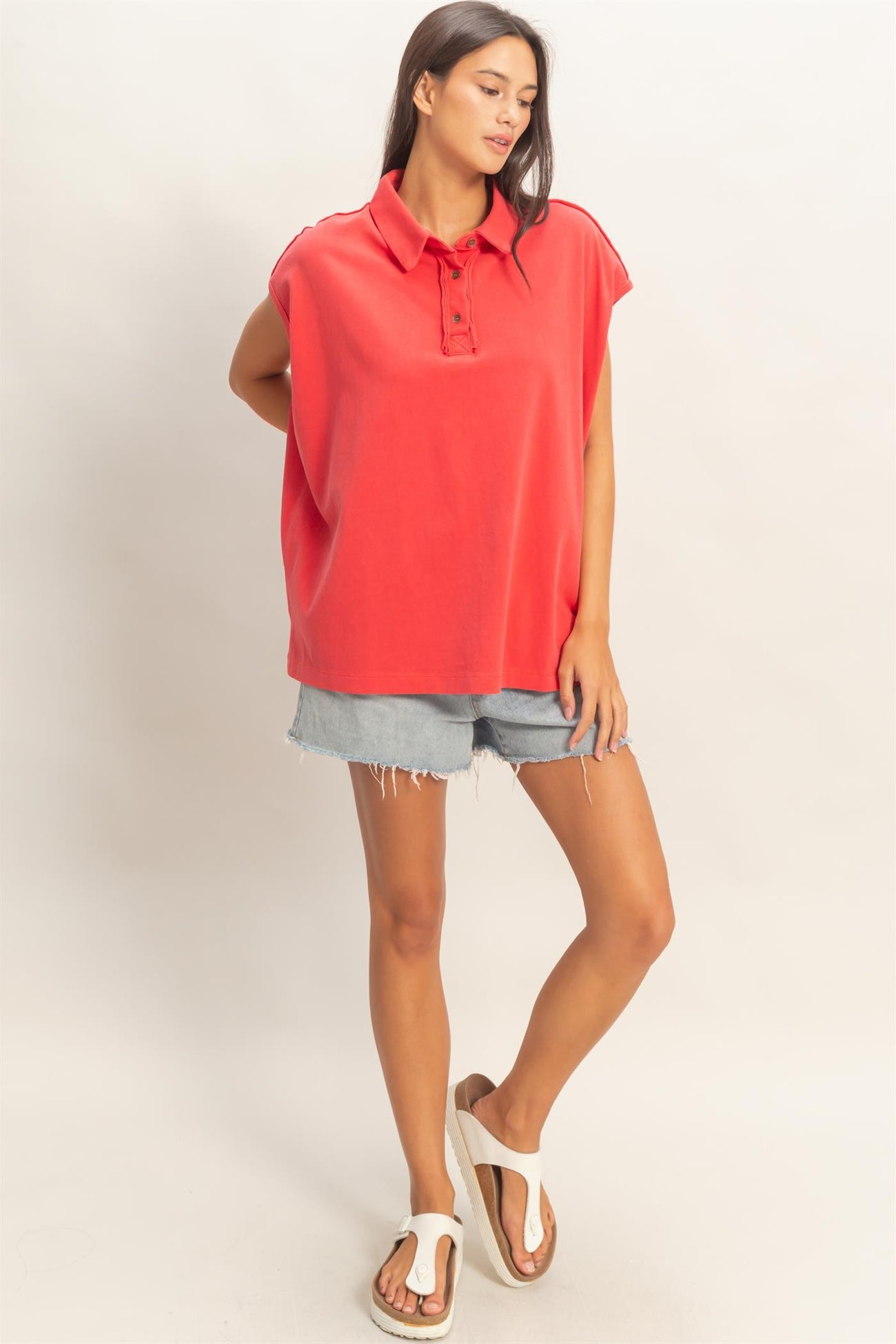 woman wearing a coral oversized polo shirt with button details and denim shorts, standing in a casual pose