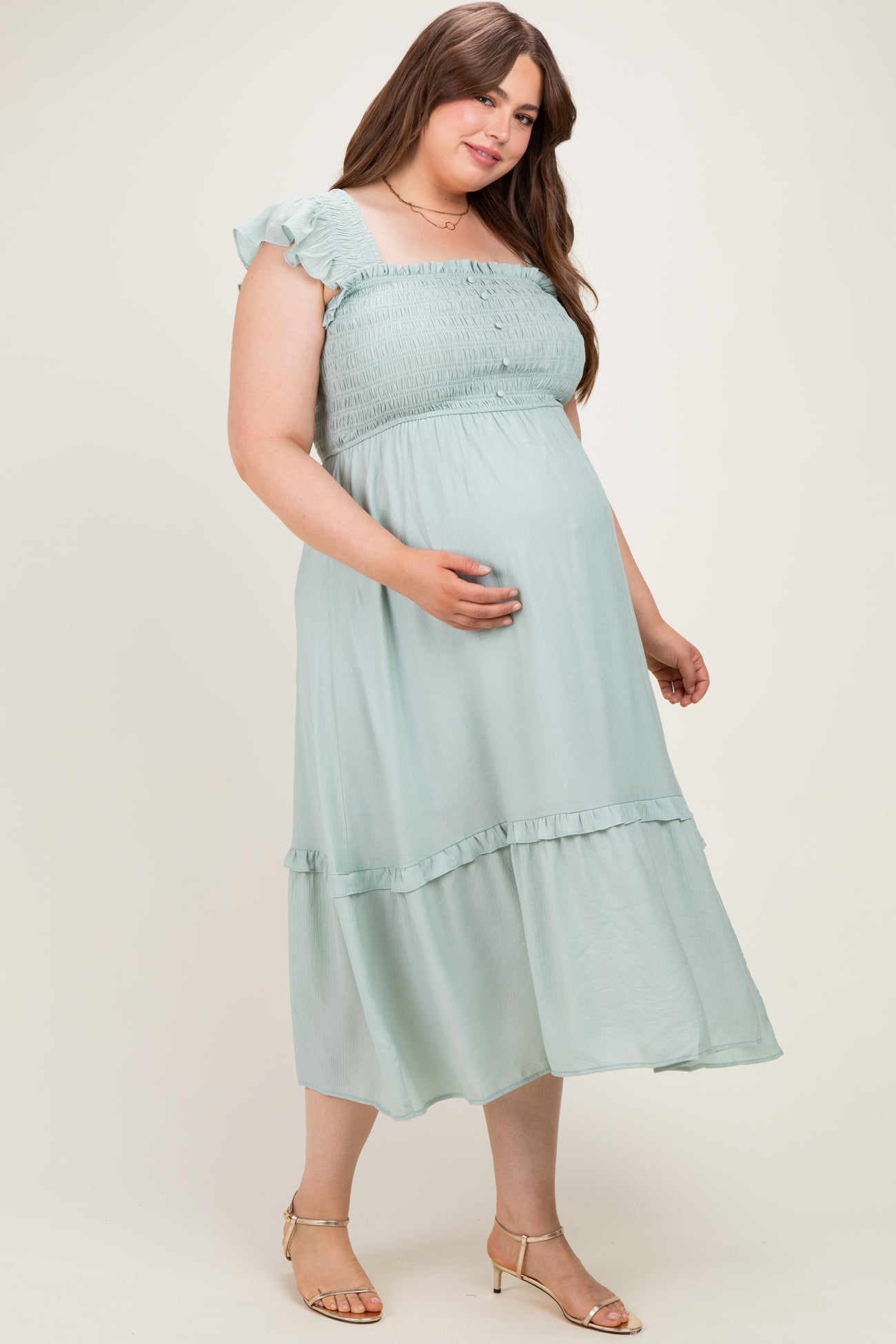 woman wearing a light green maternity plus dress with a smocked bodice and ruffled sleeves, holding her baby bump