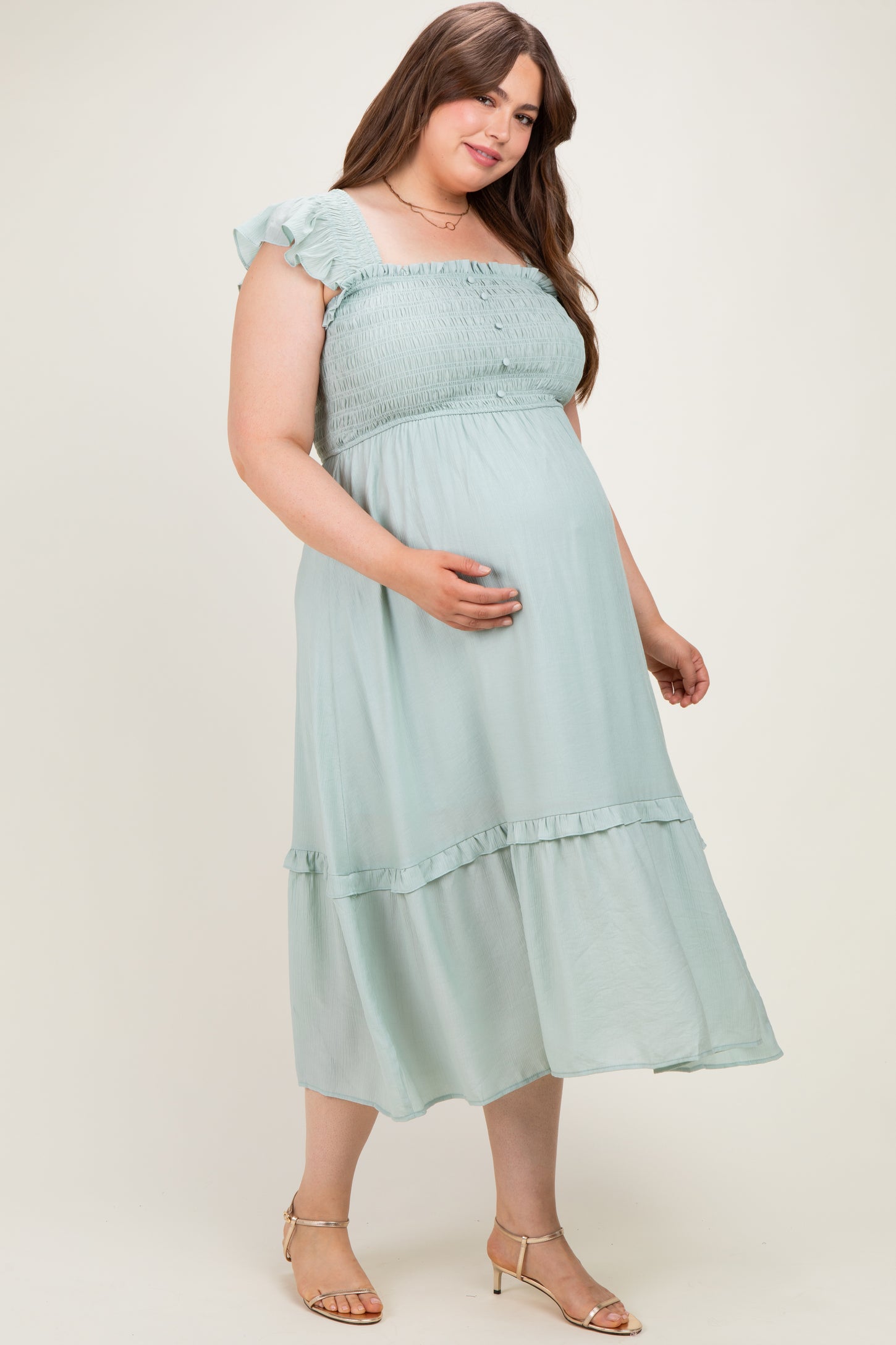 woman wearing a light green maternity plus dress with a smocked bodice and ruffled sleeves, holding her baby bump