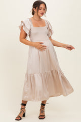 woman wearing a light beige maternity dress with ruffled sleeves and a fitted bodice, holding her baby bump