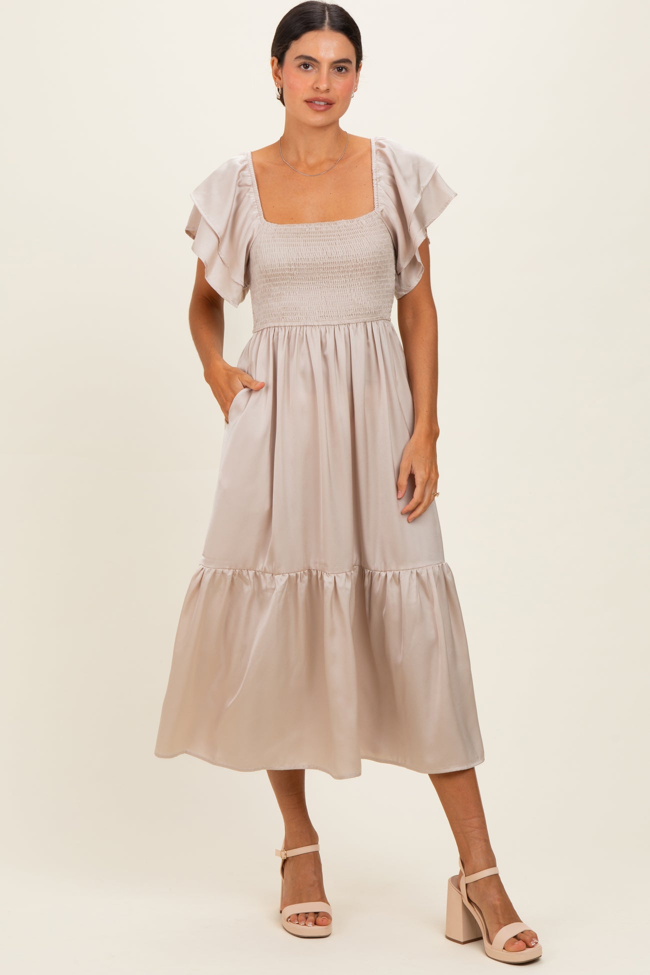 woman wearing a light beige midi dress with flutter sleeves and smocked bodice, standing with hands in pockets