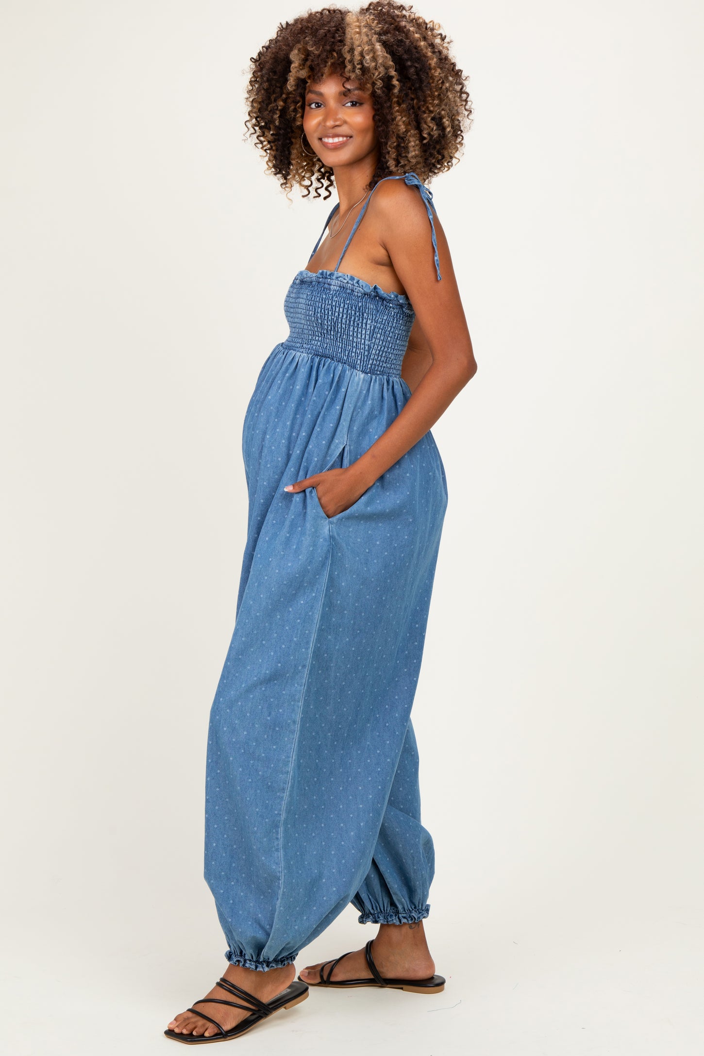 woman wearing a blue chambray maternity jumpsuit with smocked bodice and pockets, showing baby bump