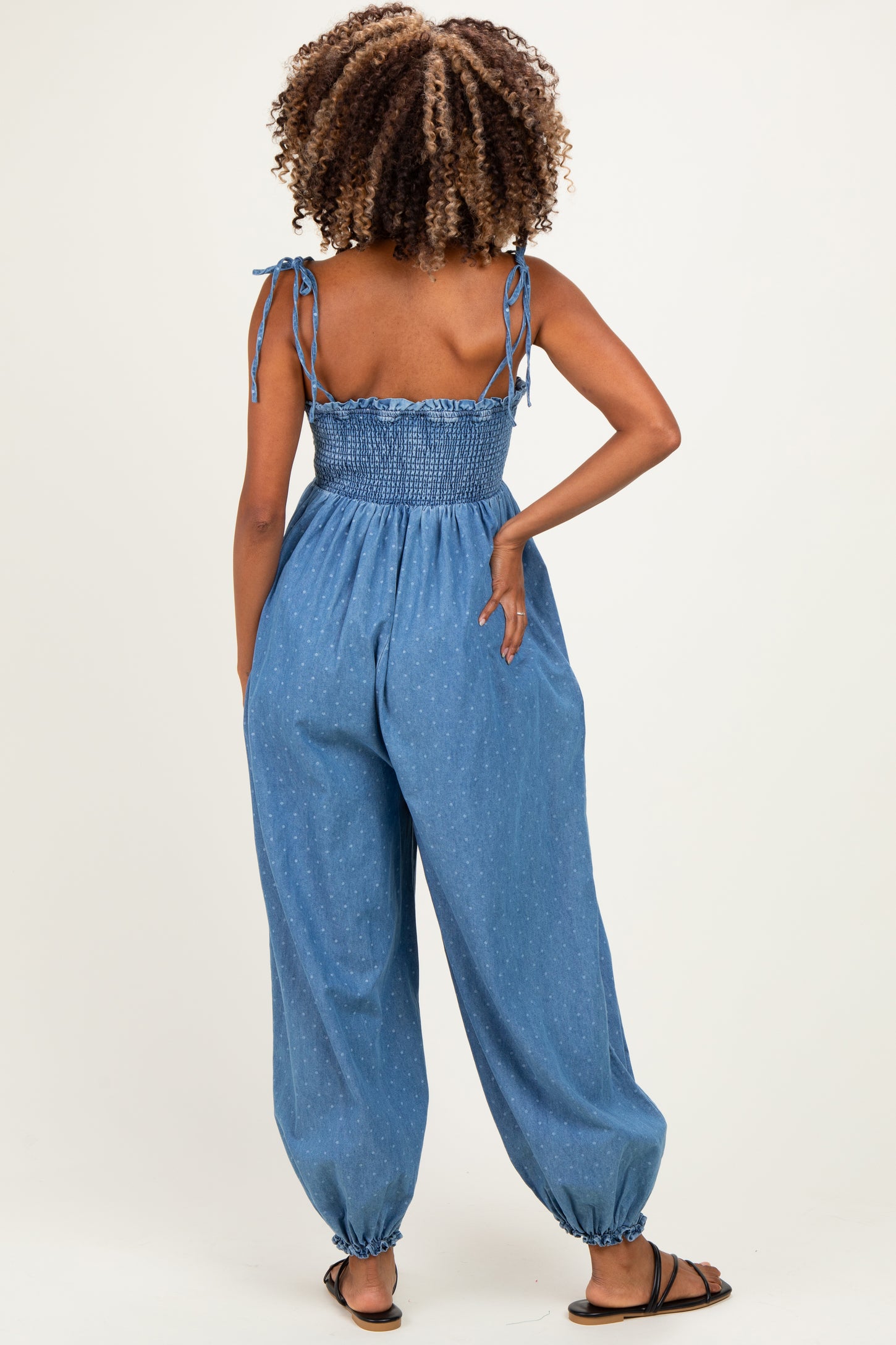 woman wearing a blue denim maternity jumpsuit with smocked bodice and tied straps, showing off her baby bump from the back
