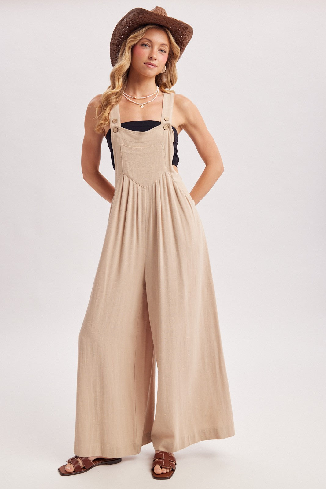woman wearing a beige wide-leg overalls with adjustable straps and a black top, styled with a hat and sandals
