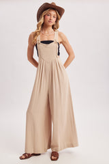 woman wearing a beige wide-leg overalls with adjustable straps and a black top, styled with a hat and sandals