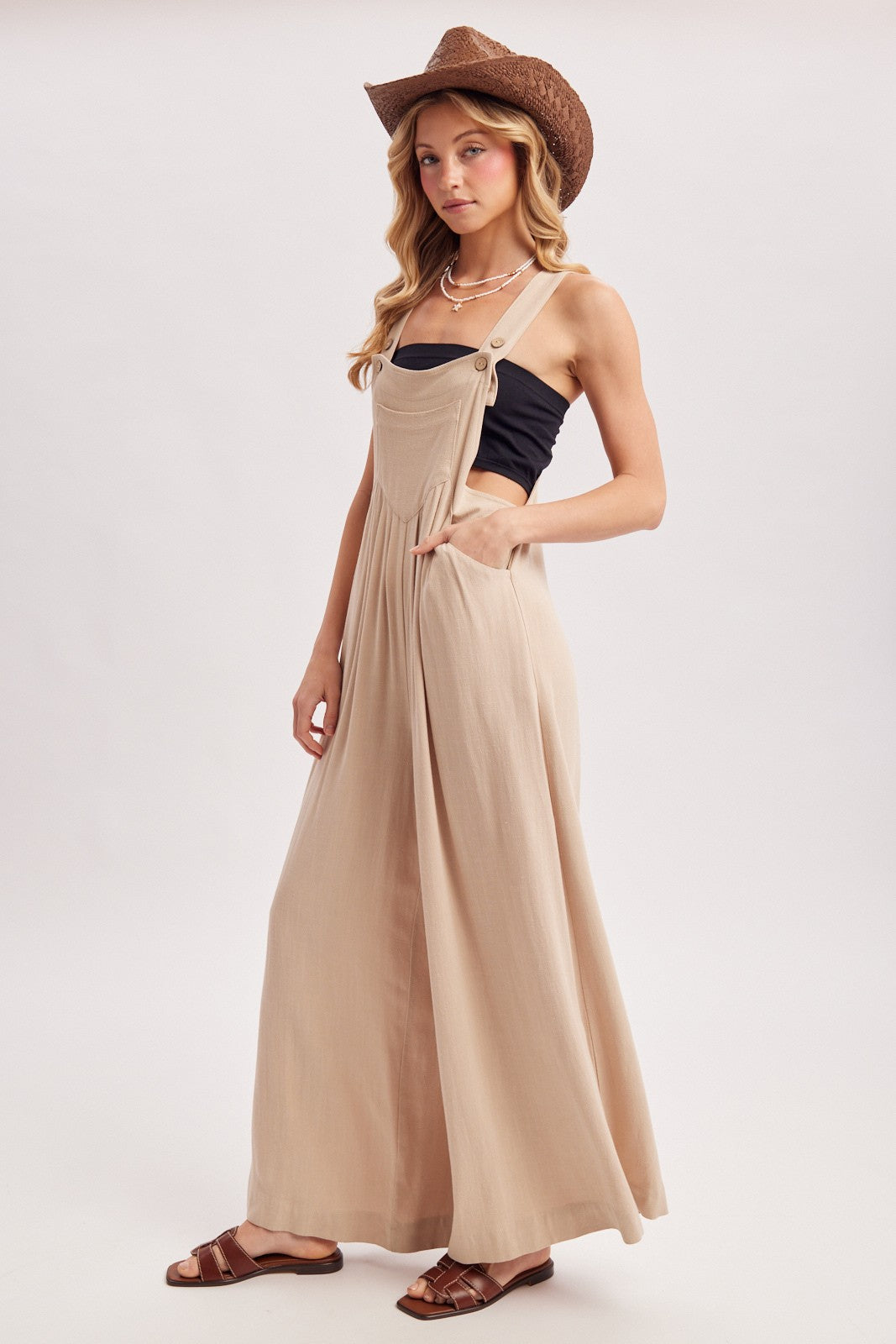 woman wearing a beige long flowy overall dress with pockets, styled with a black tank top and a brown cowboy hat