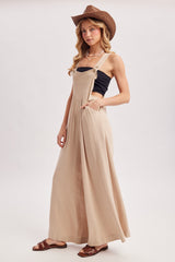 woman wearing a beige long flowy overall dress with pockets, styled with a black tank top and a brown cowboy hat