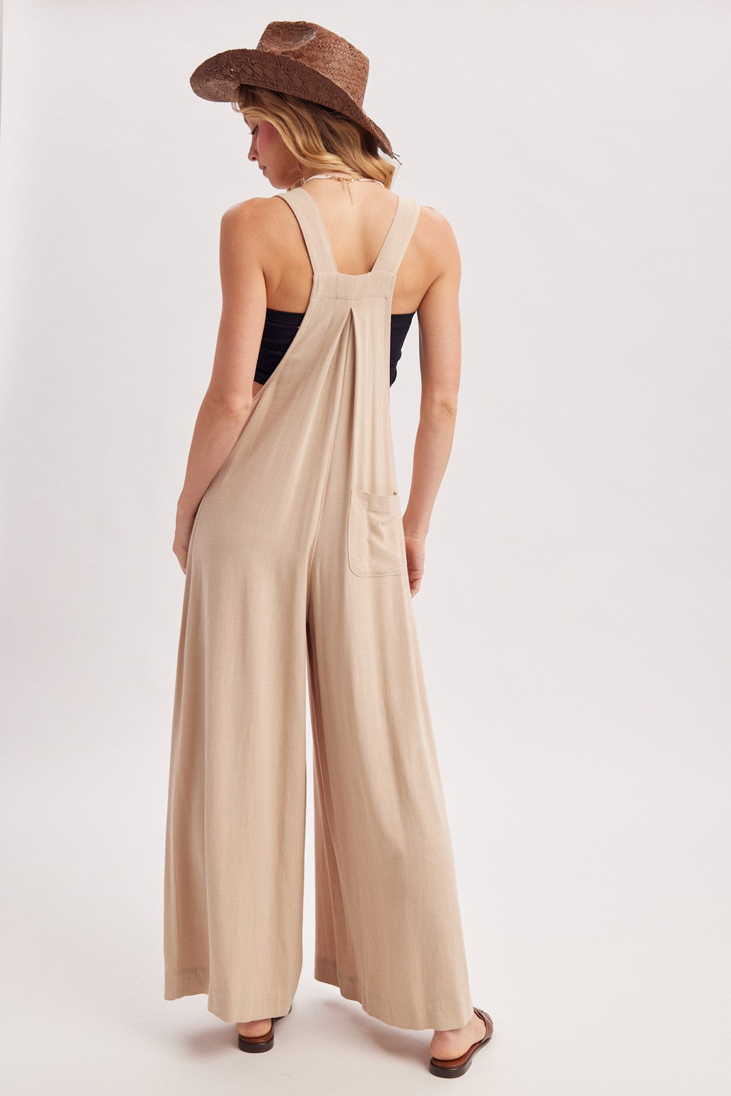 woman wearing a beige sleeveless jumpsuit with wide legs and a back pocket, styled with a straw hat