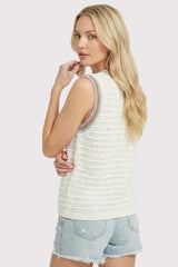 woman wearing a cream colored sleeveless sweater with a colorful stitch detail on the neckline and paired with denim shorts