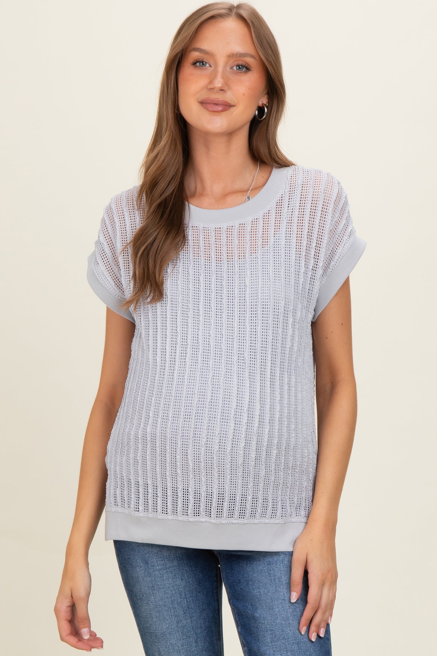 woman wearing a light grey maternity top with a sheer, mesh knit design and rolled sleeves, showing a baby bump