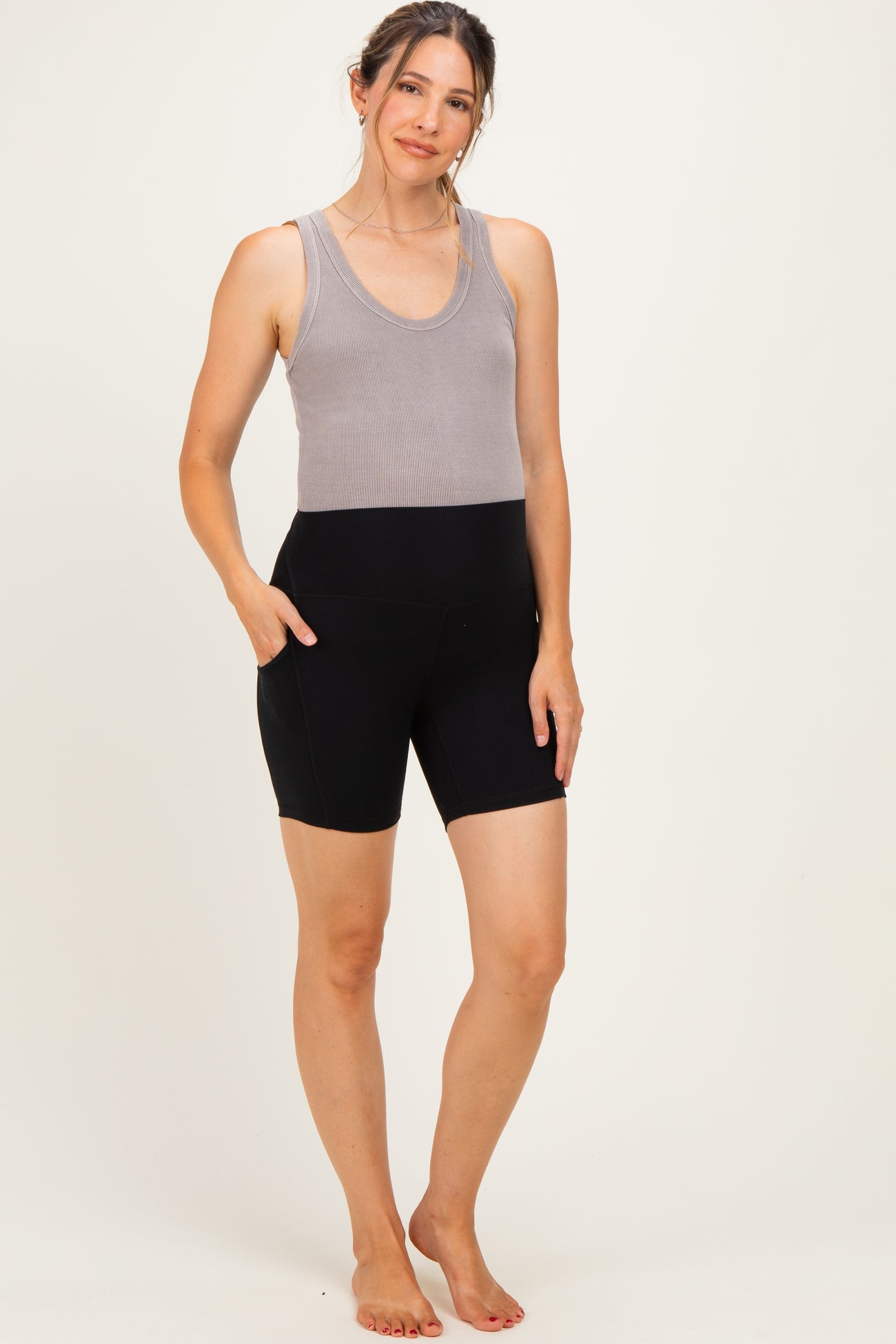 woman wearing a gray tank top and black maternity bike shorts with pockets, standing barefoot on a light background