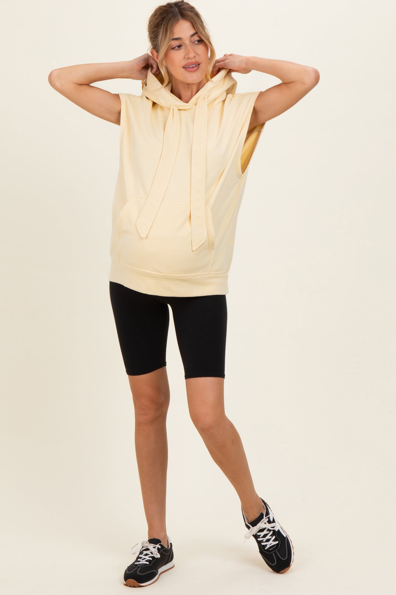 woman wearing a light yellow sleeveless maternity hoodie with a hood and black cycling shorts, showing a baby bump