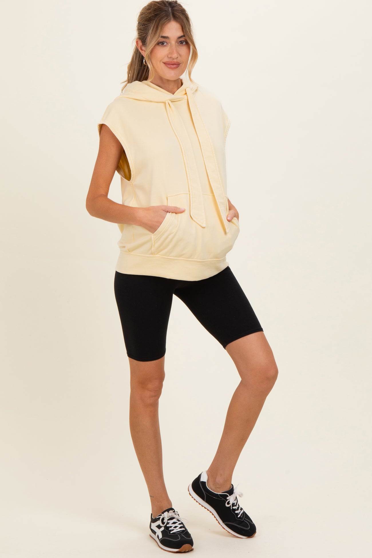woman wearing a light yellow sleeveless hoodie with pockets and black bike shorts, showcasing maternity wear