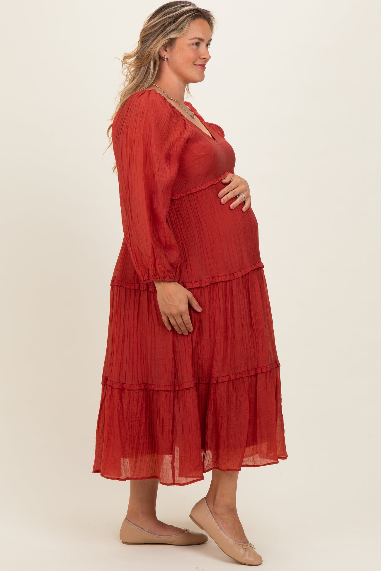woman wearing a rust red tiered maternity dress, with long sleeves and a visible baby bump, standing sideways