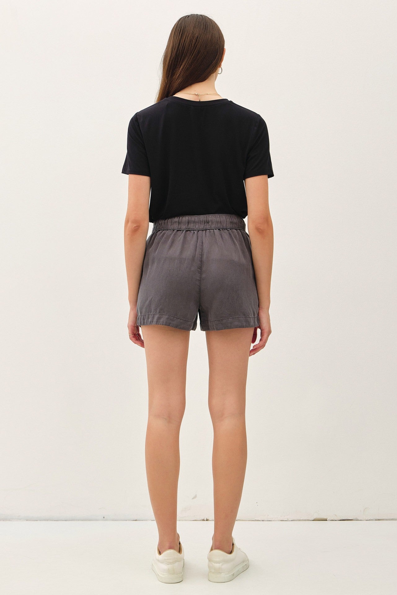 woman wearing a black t-shirt and gray shorts, viewed from the back, standing against a plain background