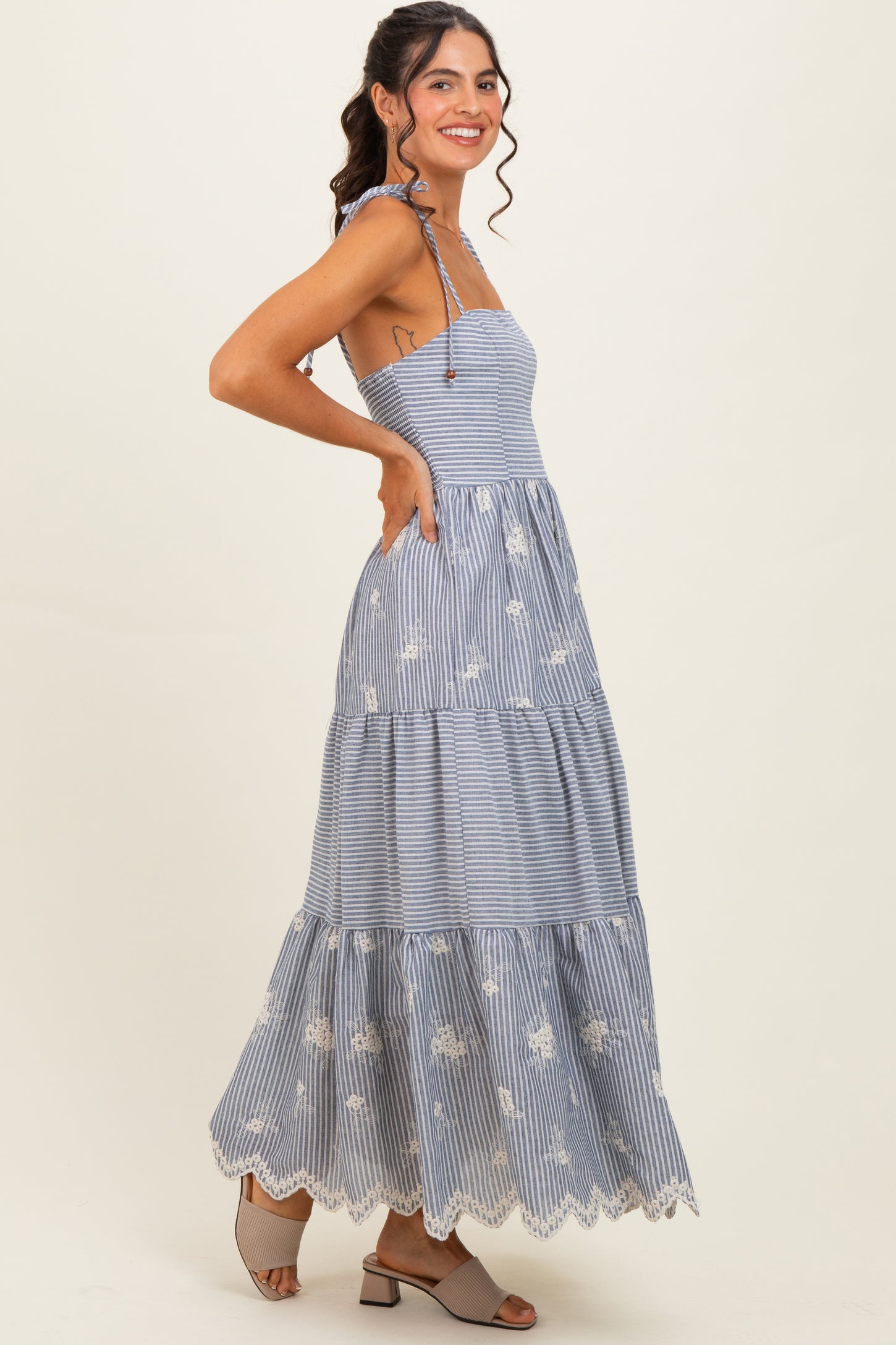 woman wearing a blue and white striped maxi dress with floral embroidery and a tiered hem, standing in profile
