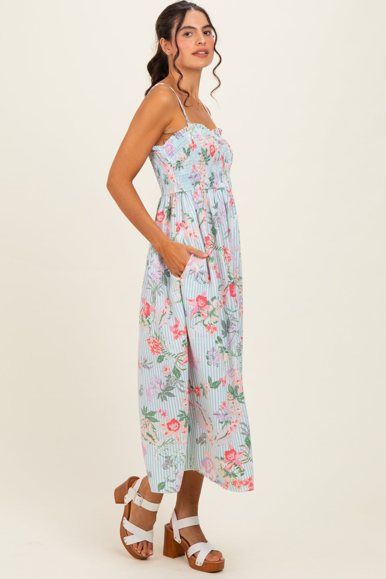 woman wearing a light blue floral maxi dress with smocked bodice and pockets, paired with white strappy sandals
