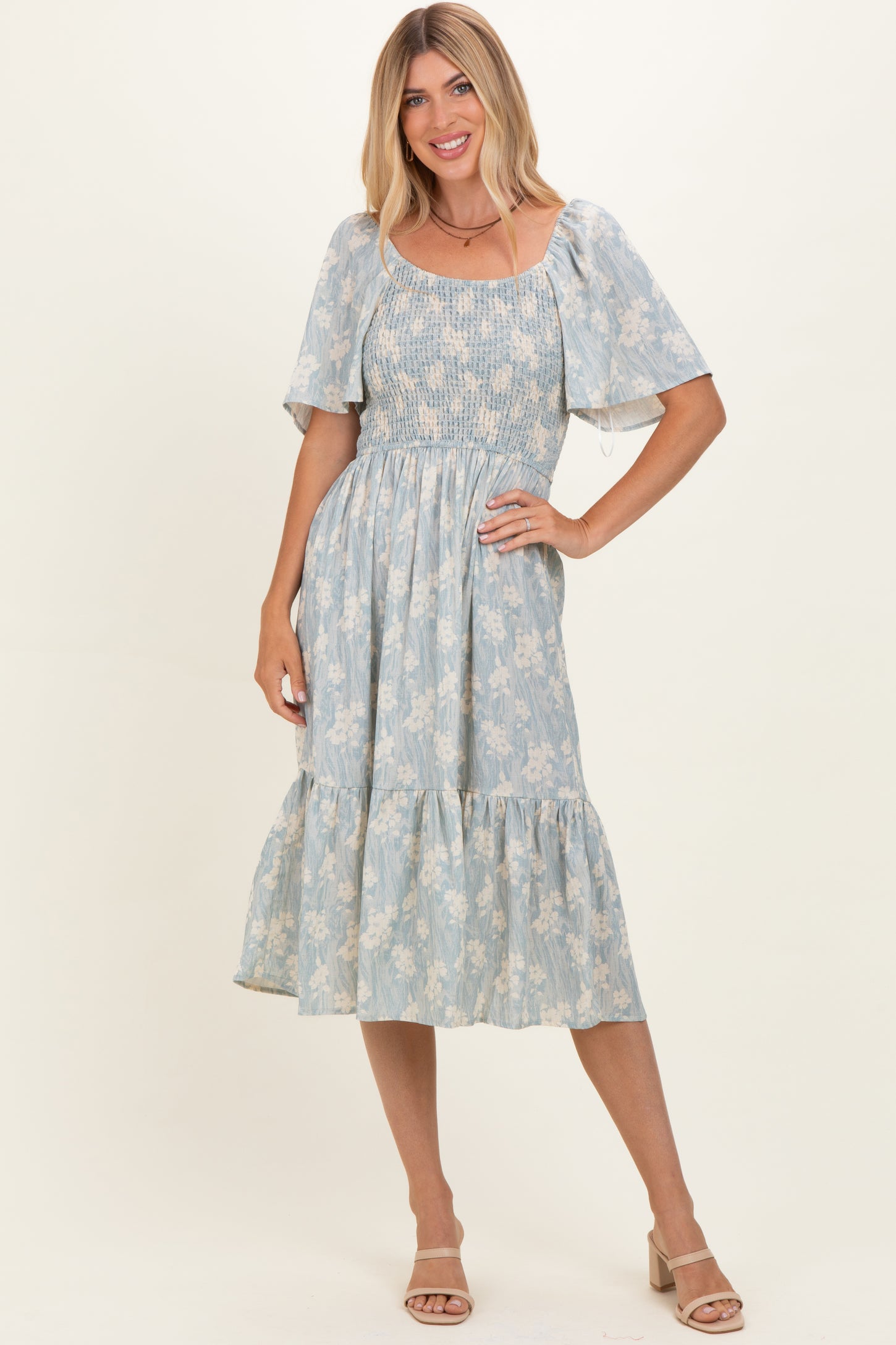 woman wearing a light blue floral midi dress with smocked bodice and flutter sleeves, standing with one hand on her hip